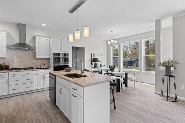 kitchen featuring wall chimney range hood, a sink, dishwasher, light wood-style flooring, and double wall oven