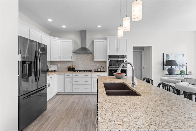 kitchen featuring white cabinetry, a sink, multiple ovens, black refrigerator with ice dispenser, and wall chimney exhaust hood