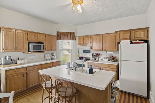 kitchen featuring white appliances, a center island with sink, light countertops, under cabinet range hood, and a sink