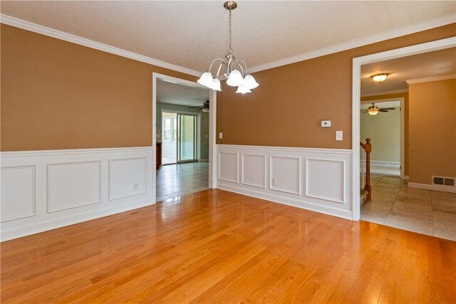 spare room with crown molding, ceiling fan with notable chandelier, a wainscoted wall, wood finished floors, and visible vents