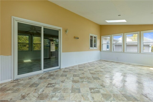 unfurnished sunroom with lofted ceiling with skylight, a wainscoted wall, stone finish floor, and a wealth of natural light