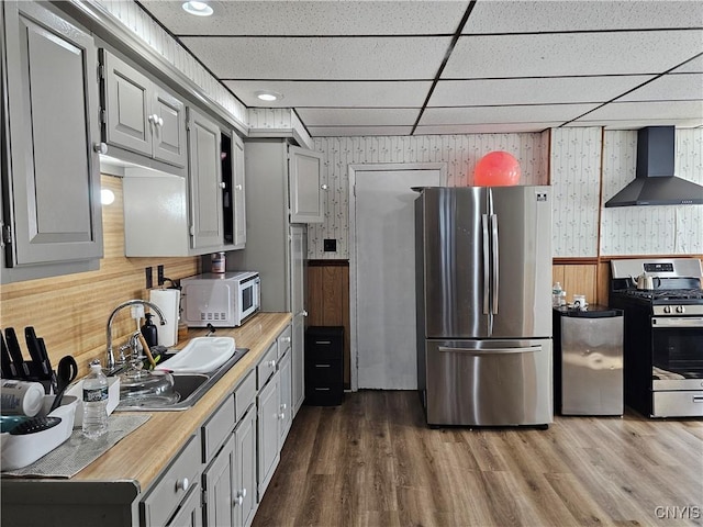 kitchen with wood finished floors, a sink, stainless steel appliances, wallpapered walls, and wall chimney exhaust hood