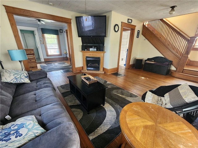living room with a ceiling fan, wood finished floors, stairway, baseboards, and a glass covered fireplace