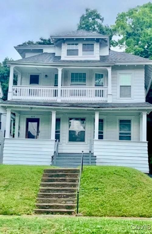 view of front facade featuring a balcony, covered porch, a front yard, roof with shingles, and stairway