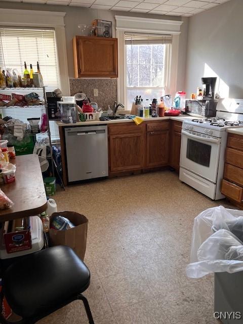 kitchen with a sink, brown cabinetry, white gas stove, a drop ceiling, and stainless steel dishwasher