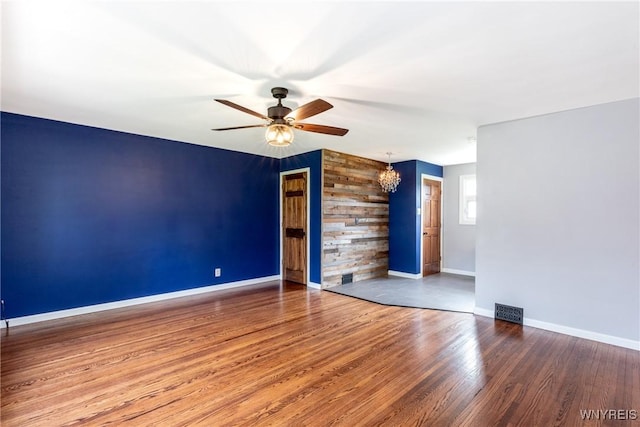 empty room featuring visible vents, an accent wall, wood finished floors, baseboards, and ceiling fan with notable chandelier