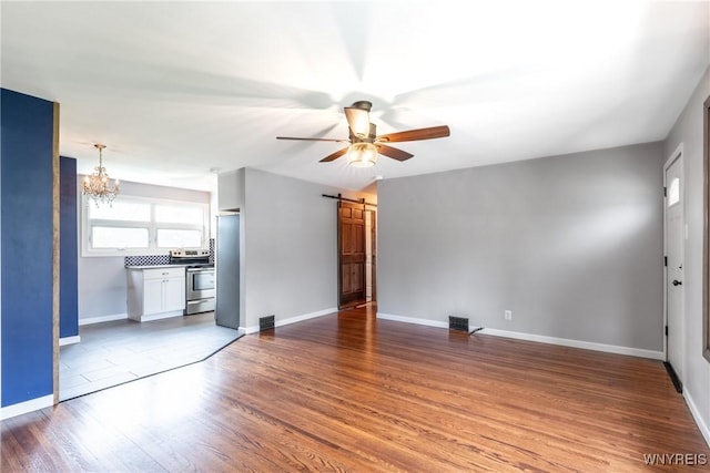 unfurnished living room featuring a barn door, ceiling fan with notable chandelier, baseboards, wood finished floors, and visible vents
