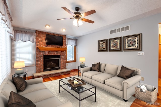 living room featuring a brick fireplace, a ceiling fan, visible vents, and wood finished floors