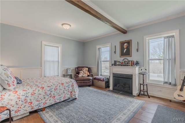 bedroom with crown molding, wood finished floors, beamed ceiling, a wainscoted wall, and a fireplace with flush hearth
