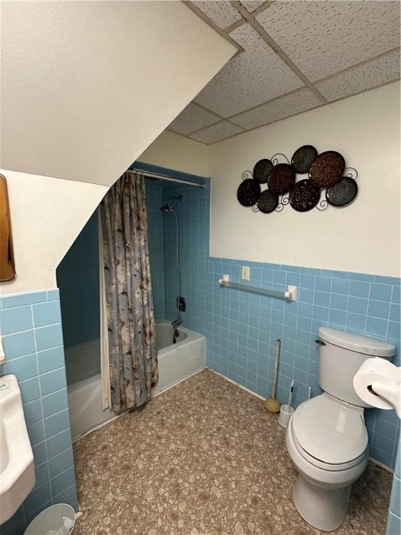 full bathroom featuring a paneled ceiling, tile walls, a wainscoted wall, toilet, and shower / bath combo with shower curtain