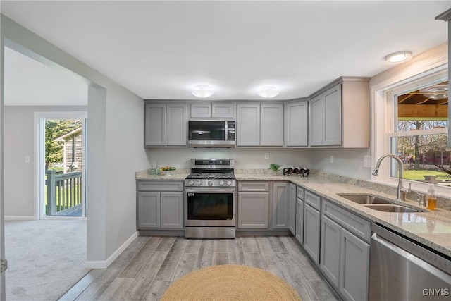 kitchen with light stone countertops, light wood-type flooring, gray cabinets, a sink, and appliances with stainless steel finishes