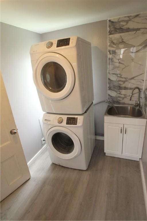 clothes washing area featuring a sink, cabinet space, wood finished floors, stacked washer and clothes dryer, and baseboards