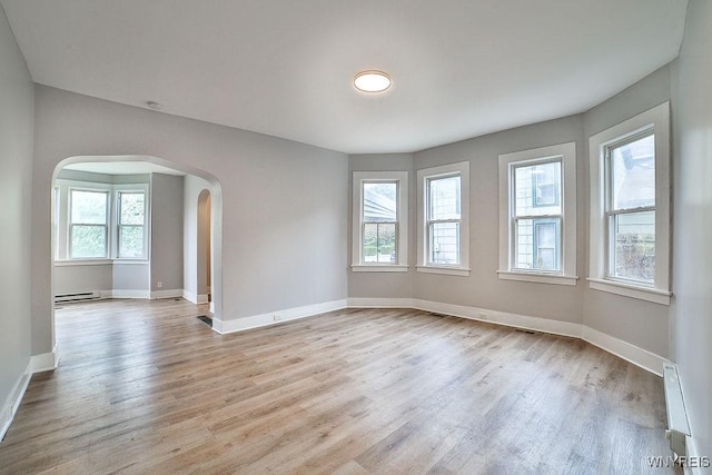 unfurnished room featuring arched walkways, a wealth of natural light, light wood finished floors, and baseboard heating