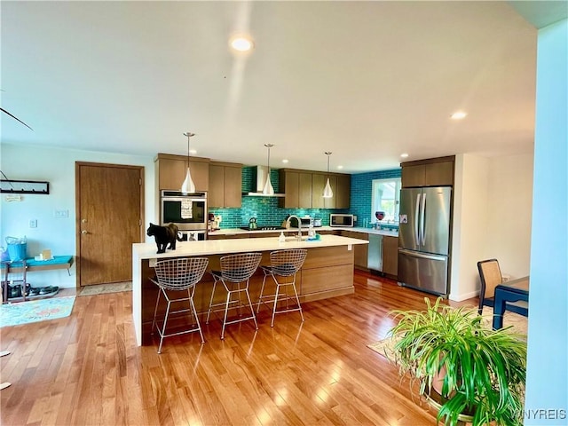 kitchen featuring light wood-style flooring, wall chimney exhaust hood, a center island with sink, light countertops, and appliances with stainless steel finishes
