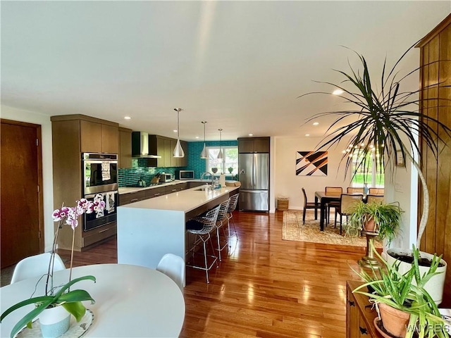kitchen with wood finished floors, a sink, wall chimney exhaust hood, stainless steel appliances, and backsplash