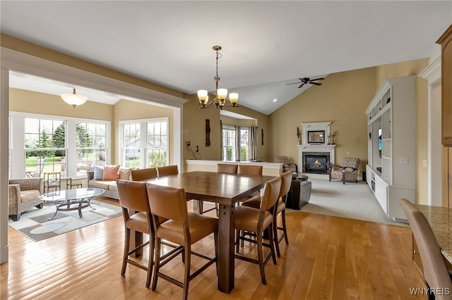 dining area with light wood-style floors, vaulted ceiling, ceiling fan with notable chandelier, and a glass covered fireplace