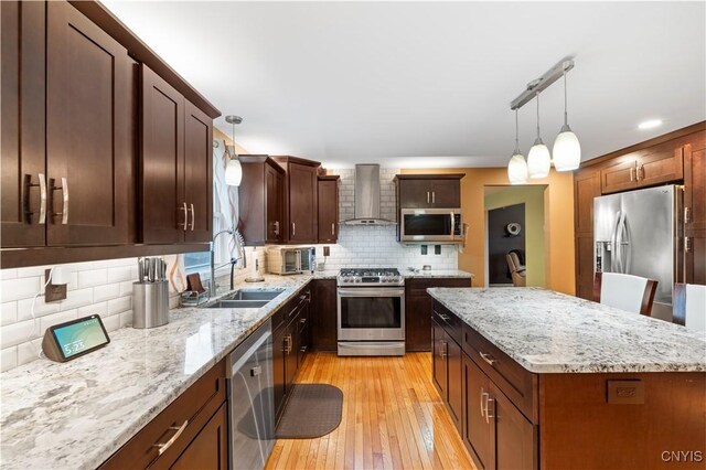 kitchen with stainless steel appliances, wall chimney range hood, a sink, light wood finished floors, and a center island