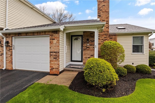 view of exterior entry with an attached garage, a chimney, aphalt driveway, a shingled roof, and brick siding