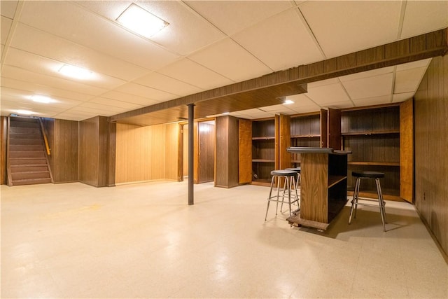 finished below grade area featuring a dry bar, wood walls, stairs, tile patterned floors, and a paneled ceiling