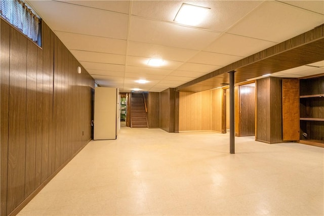 finished basement with tile patterned floors, wooden walls, stairs, and a paneled ceiling