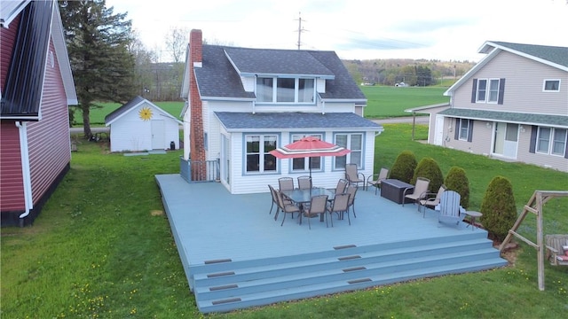 rear view of house with a shingled roof, a deck, outdoor dining space, a chimney, and a lawn