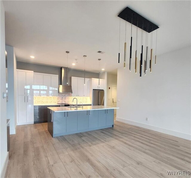 kitchen featuring wall chimney range hood, high end refrigerator, light wood-type flooring, light countertops, and a kitchen island with sink