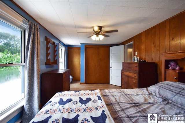 bedroom featuring multiple windows, two closets, wood finished floors, wooden walls, and a ceiling fan