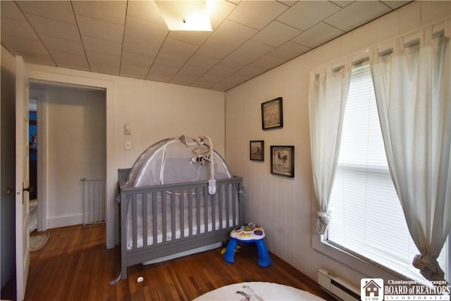 bedroom featuring a baseboard radiator, multiple windows, wood finished floors, and a nursery area