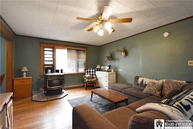 living room with a wood stove, light wood-type flooring, a ceiling fan, a baseboard heating unit, and crown molding