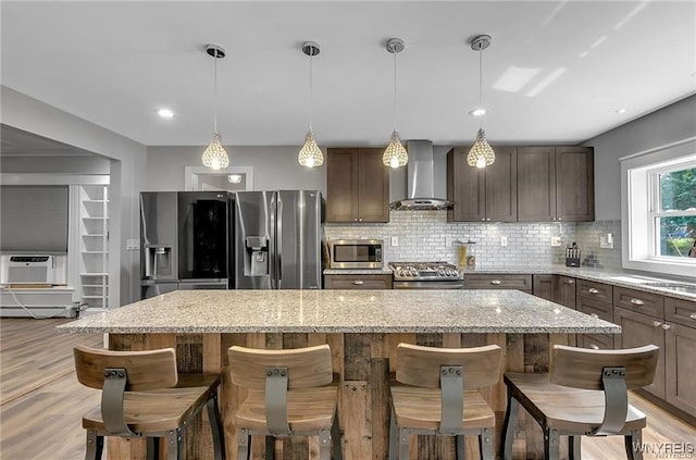 kitchen featuring wall chimney exhaust hood, appliances with stainless steel finishes, a kitchen island, backsplash, and a kitchen breakfast bar