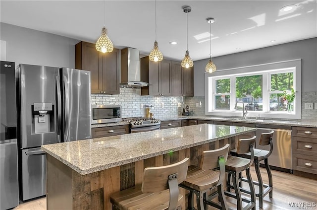 kitchen featuring wall chimney exhaust hood, stainless steel appliances, a kitchen island, backsplash, and a kitchen breakfast bar