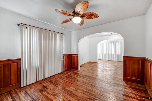 unfurnished room featuring a wainscoted wall, wood finished floors, a ceiling fan, and arched walkways