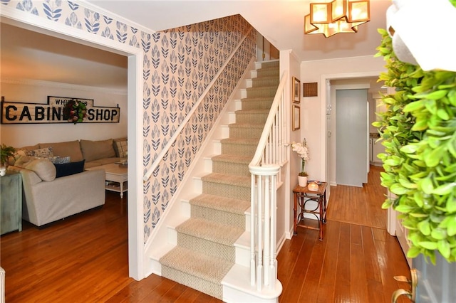 staircase featuring wallpapered walls, wood-type flooring, ornamental molding, baseboards, and a chandelier