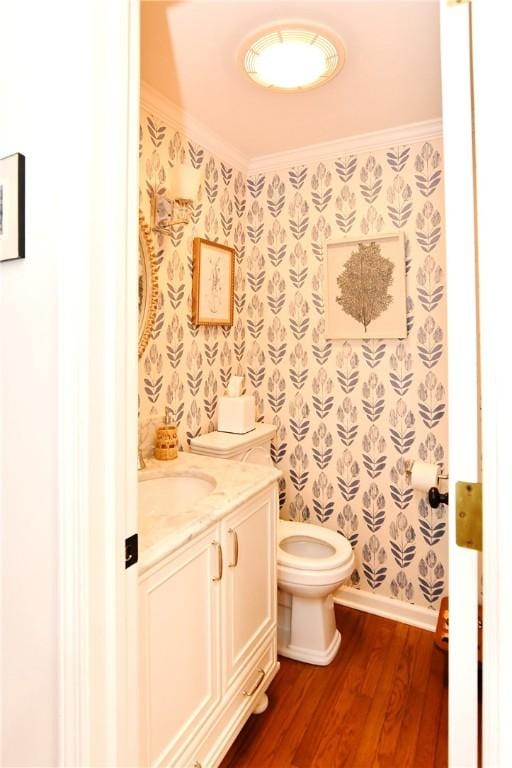 half bathroom with wood finished floors, crown molding, vanity, toilet, and wallpapered walls