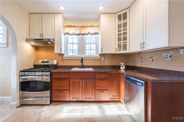 kitchen with appliances with stainless steel finishes, under cabinet range hood, a sink, arched walkways, and dark countertops