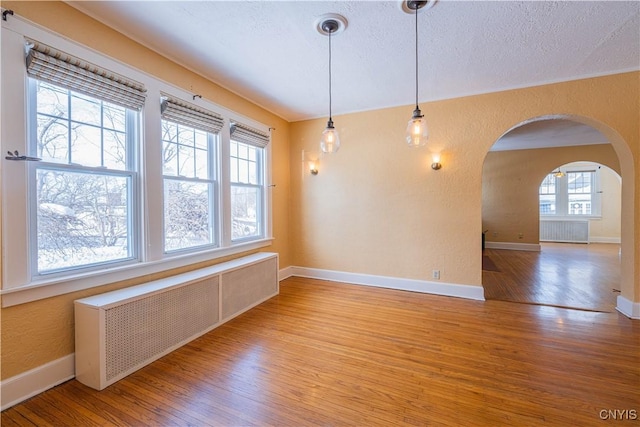 unfurnished room featuring radiator heating unit, wood finished floors, arched walkways, baseboards, and a textured wall