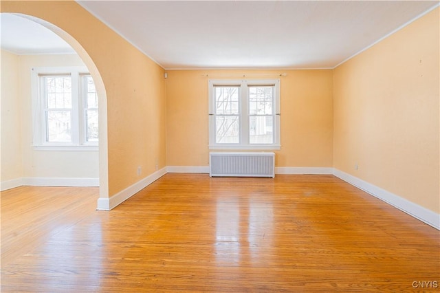 empty room with radiator, light wood-style flooring, arched walkways, crown molding, and baseboards