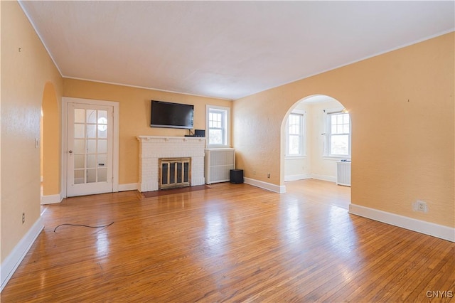 unfurnished living room featuring arched walkways, wood finished floors, a brick fireplace, baseboards, and crown molding