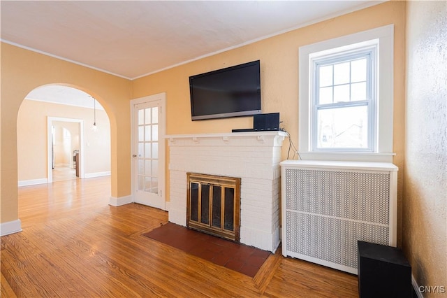unfurnished living room with wood finished floors, radiator heating unit, crown molding, a brick fireplace, and arched walkways