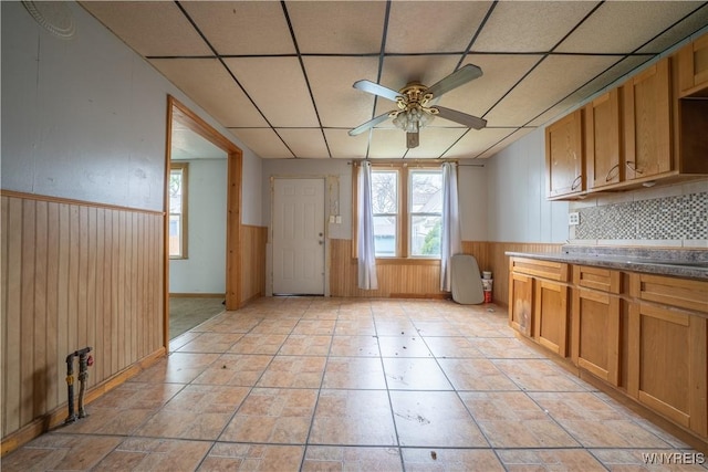 kitchen featuring a ceiling fan, a paneled ceiling, wainscoting, brown cabinetry, and wood walls