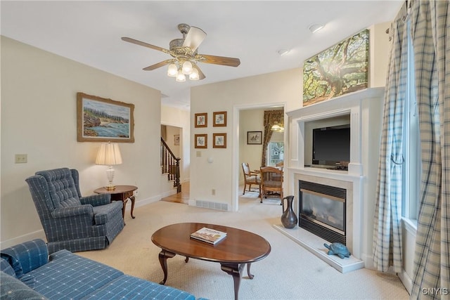 living area featuring stairs, light colored carpet, ceiling fan, a glass covered fireplace, and baseboards