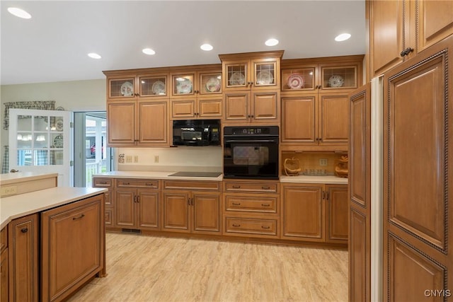 kitchen featuring black appliances, brown cabinetry, light countertops, light wood-type flooring, and glass insert cabinets