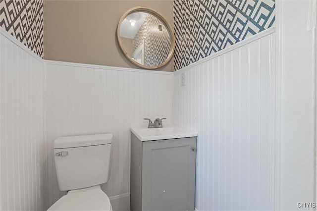 half bathroom with a wainscoted wall, toilet, vanity, and wallpapered walls