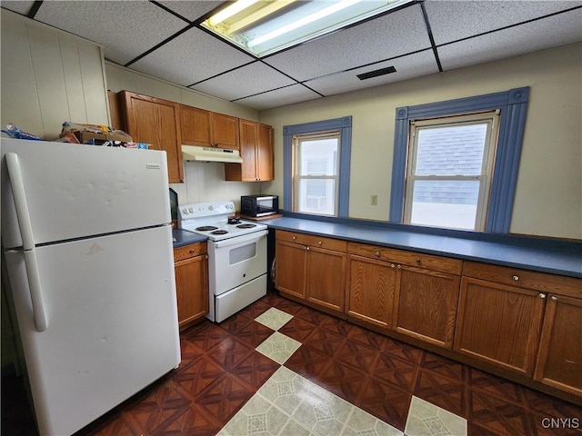 kitchen with white appliances, under cabinet range hood, a drop ceiling, brown cabinets, and dark countertops