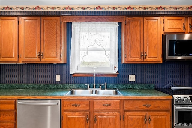 kitchen featuring appliances with stainless steel finishes, a sink, wallpapered walls, brown cabinets, and dark countertops