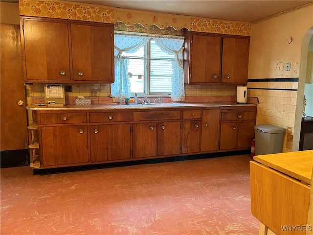 kitchen with brown cabinets, arched walkways, a sink, light flooring, and tile walls