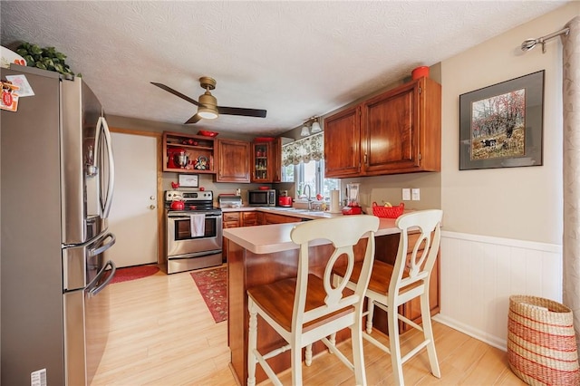 kitchen with stainless steel appliances, a peninsula, ceiling fan, light wood-type flooring, and a wainscoted wall