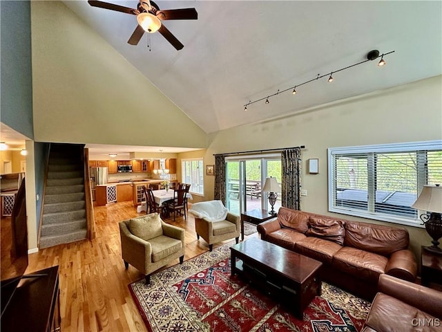 living room featuring high vaulted ceiling, a ceiling fan, stairway, and light wood-style flooring