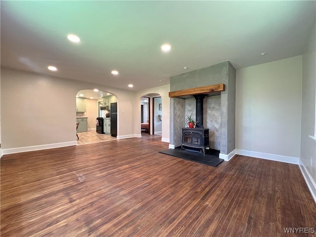 unfurnished living room with arched walkways, wood finished floors, recessed lighting, baseboards, and a wood stove