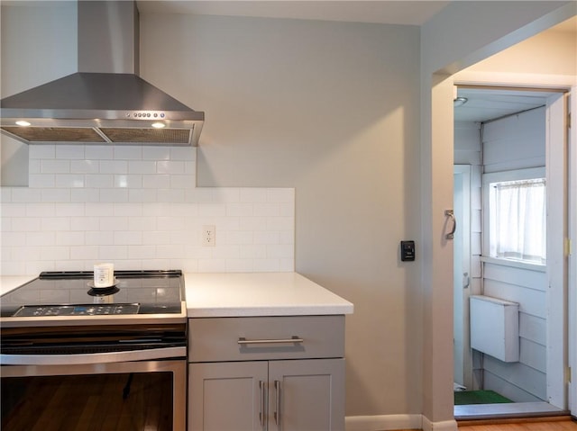 kitchen featuring electric range, wall chimney range hood, gray cabinets, backsplash, and light countertops